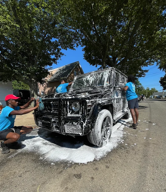 New Era staff foam-washing a black Mercedes G-Class at the curbside
